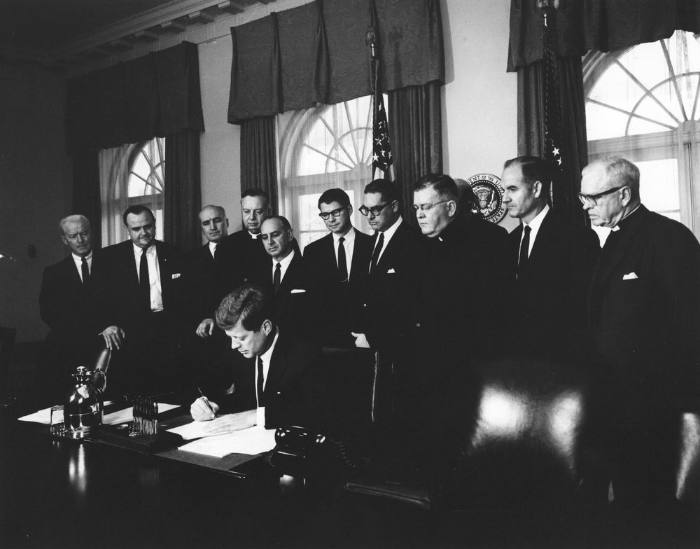 Flanked by officials representing several ACVAFS members, JFK signs a proclamation designating the week of April 9, 1962, as Voluntary Overseas Aid Week. Source: http://www.jfklibrary.org/Asset-Viewer/Archives/JFKWHP-AR7157-D.aspx